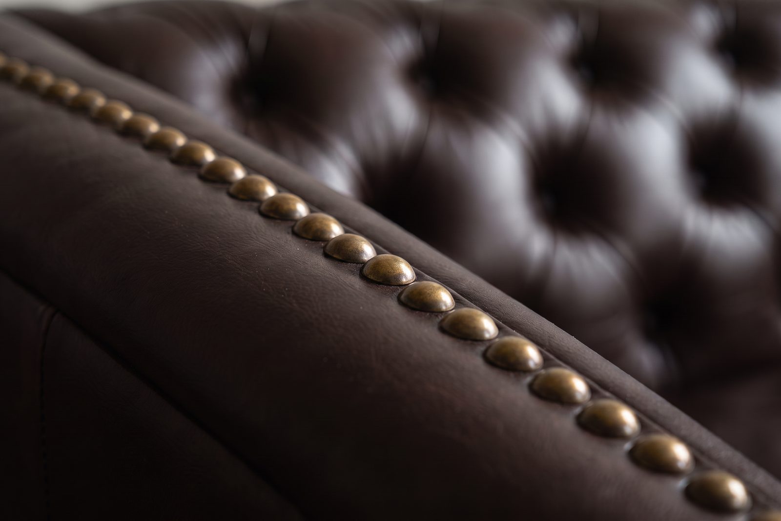 Row of antique-brass nailhead studs running along the rolled arm of a dark brown leather Chesterfield, each individually placed