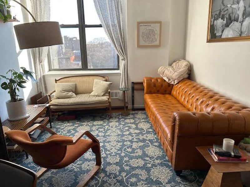 Customer photograph: a tan buttoned leather Chesterfield in a bright apartment living room, with a patterned blue rug and a bentwood chair