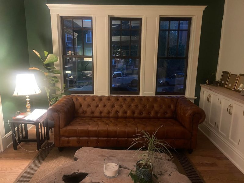 Customer photograph: a tan buttoned leather Chesterfield against a deep green wall beneath three sash windows, with a live-edge coffee table